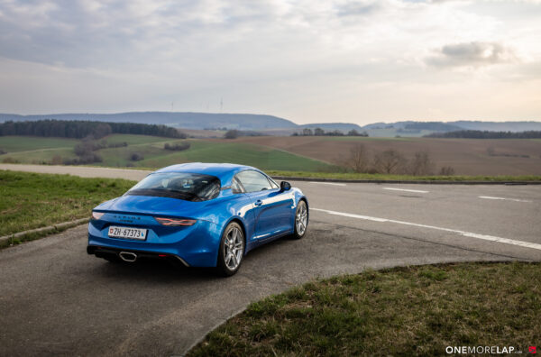 Blue Alpine sports car parked on a rural road with green fields and rolling hills in the background.
